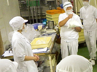 パン製造技術講習会で、白衣姿の参加者が製パン実習中に講師から指導を受けている様子。