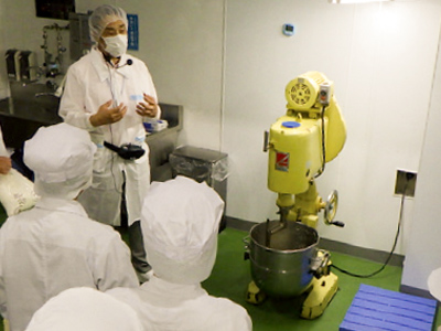 製パン実習室で講師が大型ミキサーの前で説明を行い、参加者が周囲で話を聞いている。