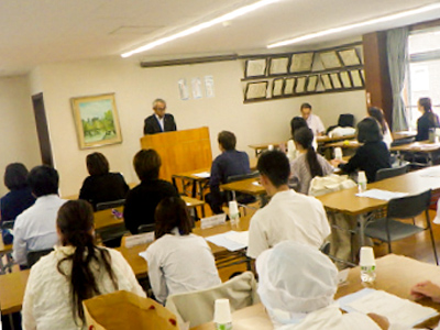 講習会の座学風景。講師が演台に立ち、受講者がノートを取りながら説明を聞いている。