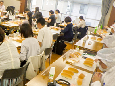 パン製造技術講習会の試食・実習風景。参加者が机に並んだパンを前に講師の説明を聞いている。