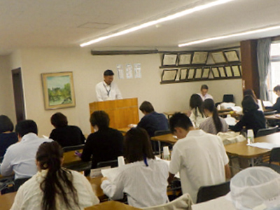 講習会の座学の様子。講師が演台で説明を行い、受講者が整然と並んで講義に参加している。